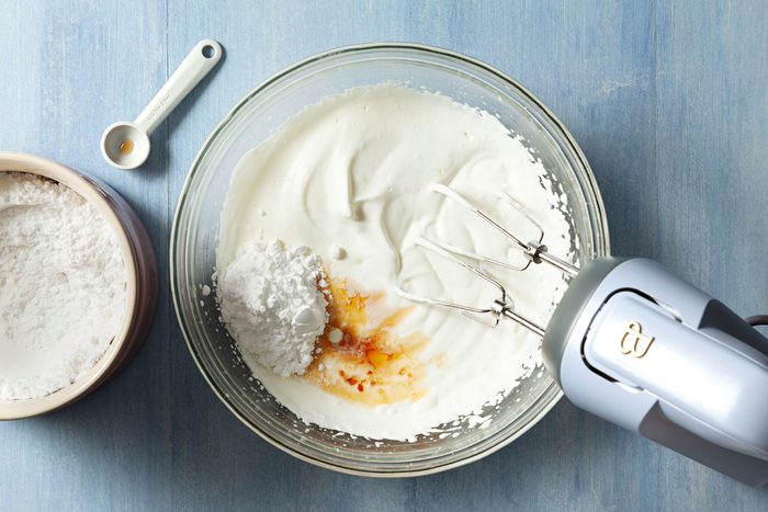 beating cream, sugar and vanilla In a large glass bowl using a hand mixer on a textured light blue surface