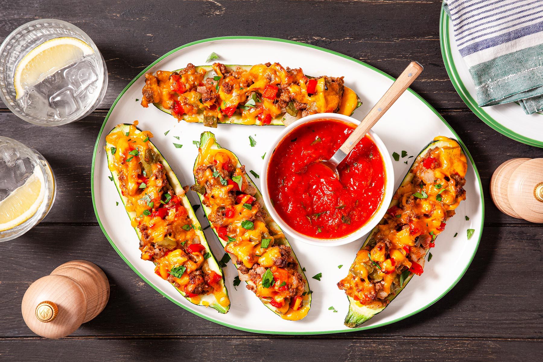 Overhead shot of Zucchini Boats; sauce; salt shaker; plates; napkin; glass drinks; wooden background;