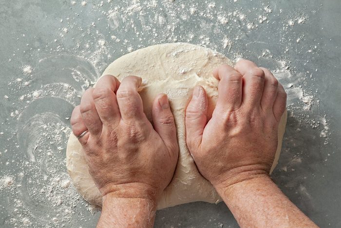 Two hands kneading a ball of dough on a floured surface. Flour is scattered around, indicating the process of preparing the dough.