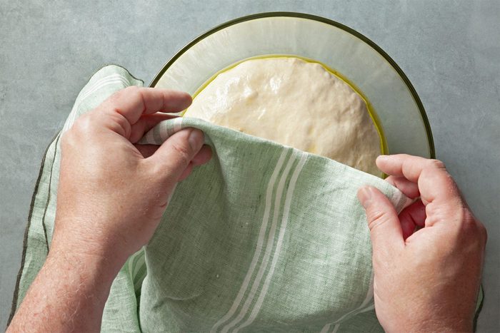 A pair of hands is seen lifting a green cloth to reveal a bowl containing risen dough. The bowl sits on a gray surface, and the dough has a light, smooth texture.