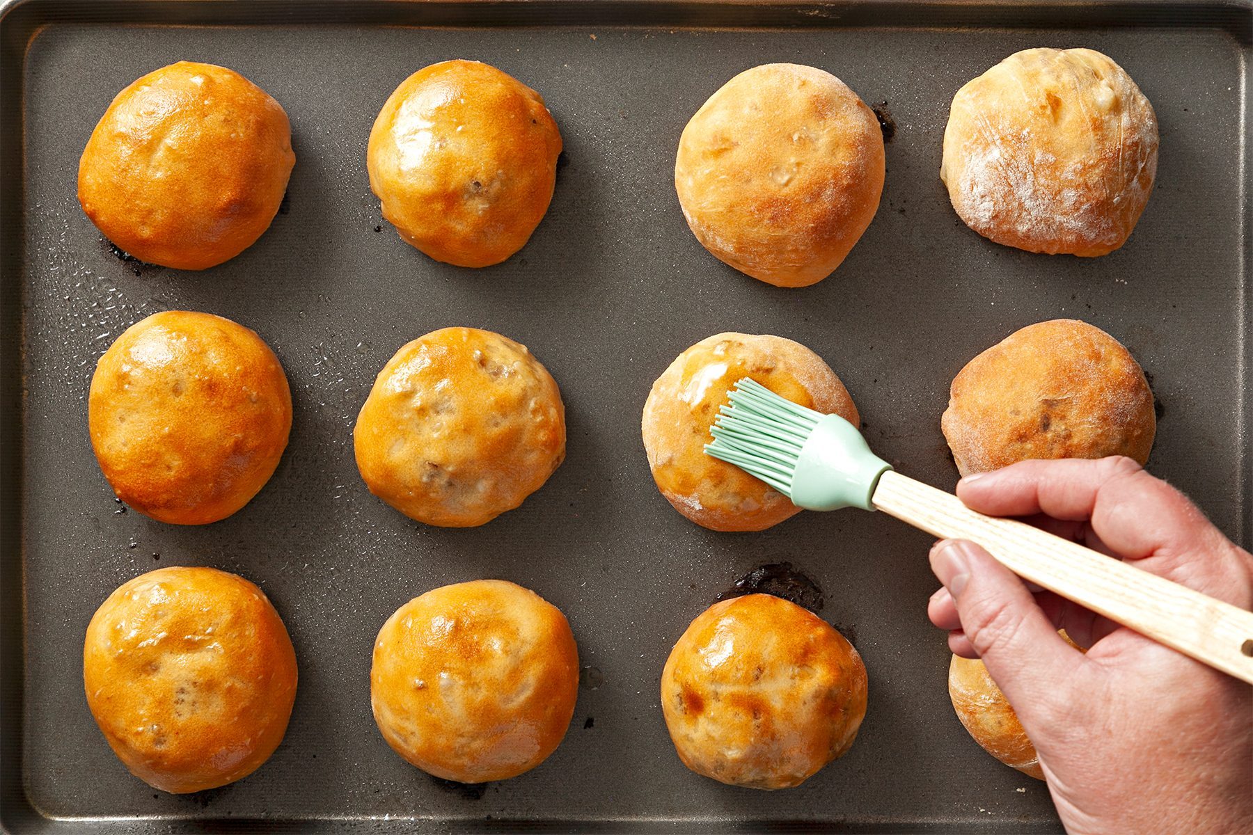 A hand uses a brush to glaze golden brown dinner rolls on a baking sheet. The rolls, neatly arranged in a 4x3 grid, have a shiny, appetizing appearance, indicating they are freshly baked.