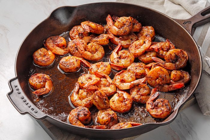 High angle shot of a large cast iron skillet; melt butter over medium heat; added shrimp; cook and stir until shrimp turn pink and edges are browned; wooden base; kitchen towel; white marble background;