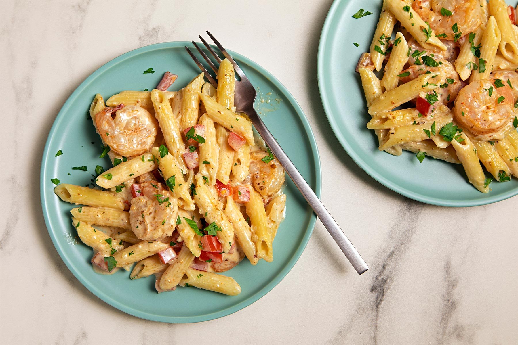 Two plates of creamy penne pasta with shrimp are on a marble surface. The pasta is garnished with chopped parsley and includes pieces of red onion and diced tomatoes. A fork is placed on one of the plates.