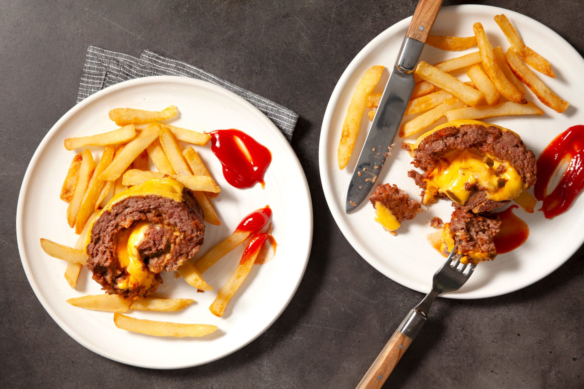 Overhead shot of Cheeseburger Meat Loaf; served on two white plates; with fries and ketchup; cut into small round pieces; cutlery; white and black lined napkin cloth; black texture background;
