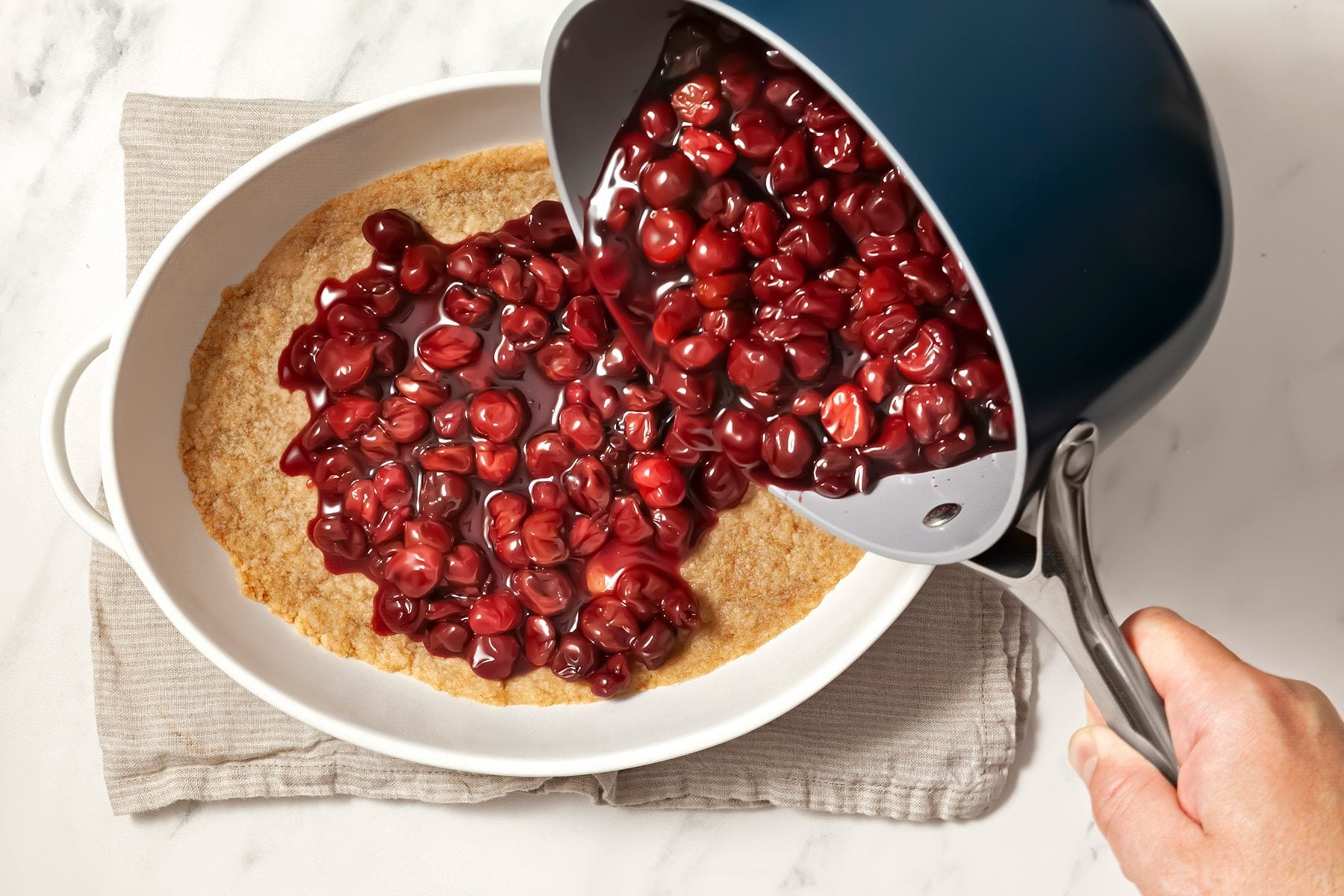 3/4th shot; white background; pouring cherries mixture from saucepan over baked crust placed over kitchen towel