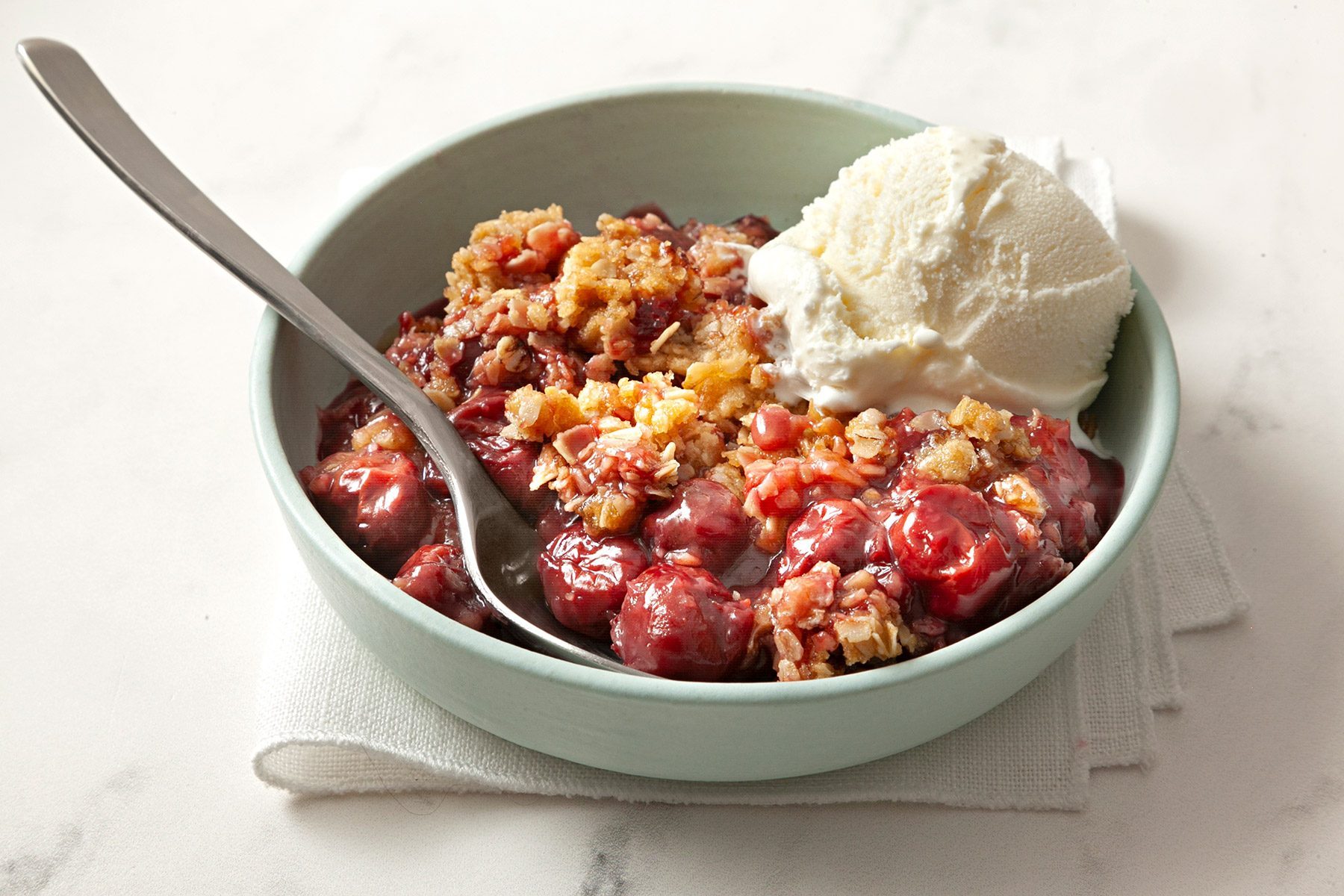 3/4th shot; white background; Cherry Crisp served in white bowl over kitchen napkin with ice cream and silver spoon