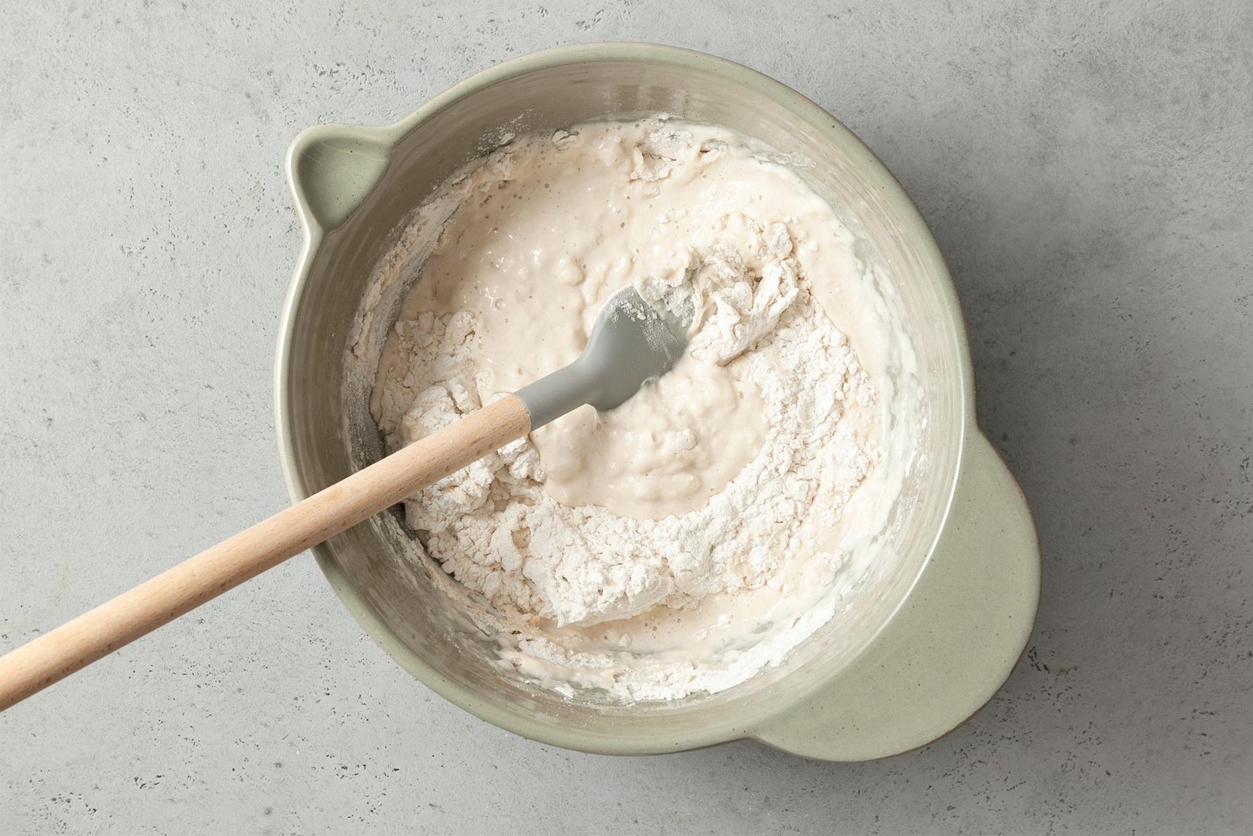 A light green mixing bowl filled with partially mixed dough, with visible dry flour and wet mixture. A gray silicone spatula with a wooden handle is placed inside the bowl, resting on the edge. 