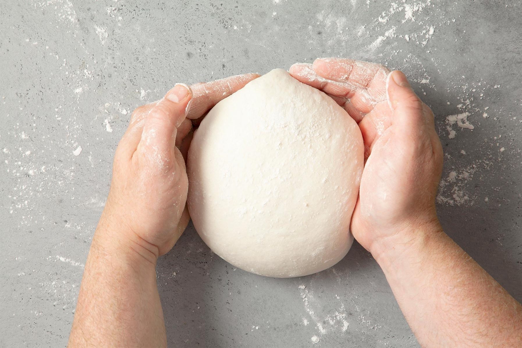 Two hands, lightly dusted with flour, are shaping a round, smooth ball of dough on a floured grey countertop. 