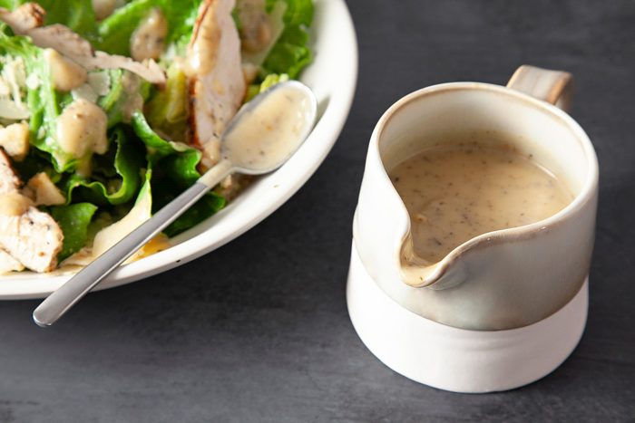 3/4 angle view shot of Roasted Garlic Vinaigrette; in a ceramic jar; dressing over salad; spoon; dark grey background