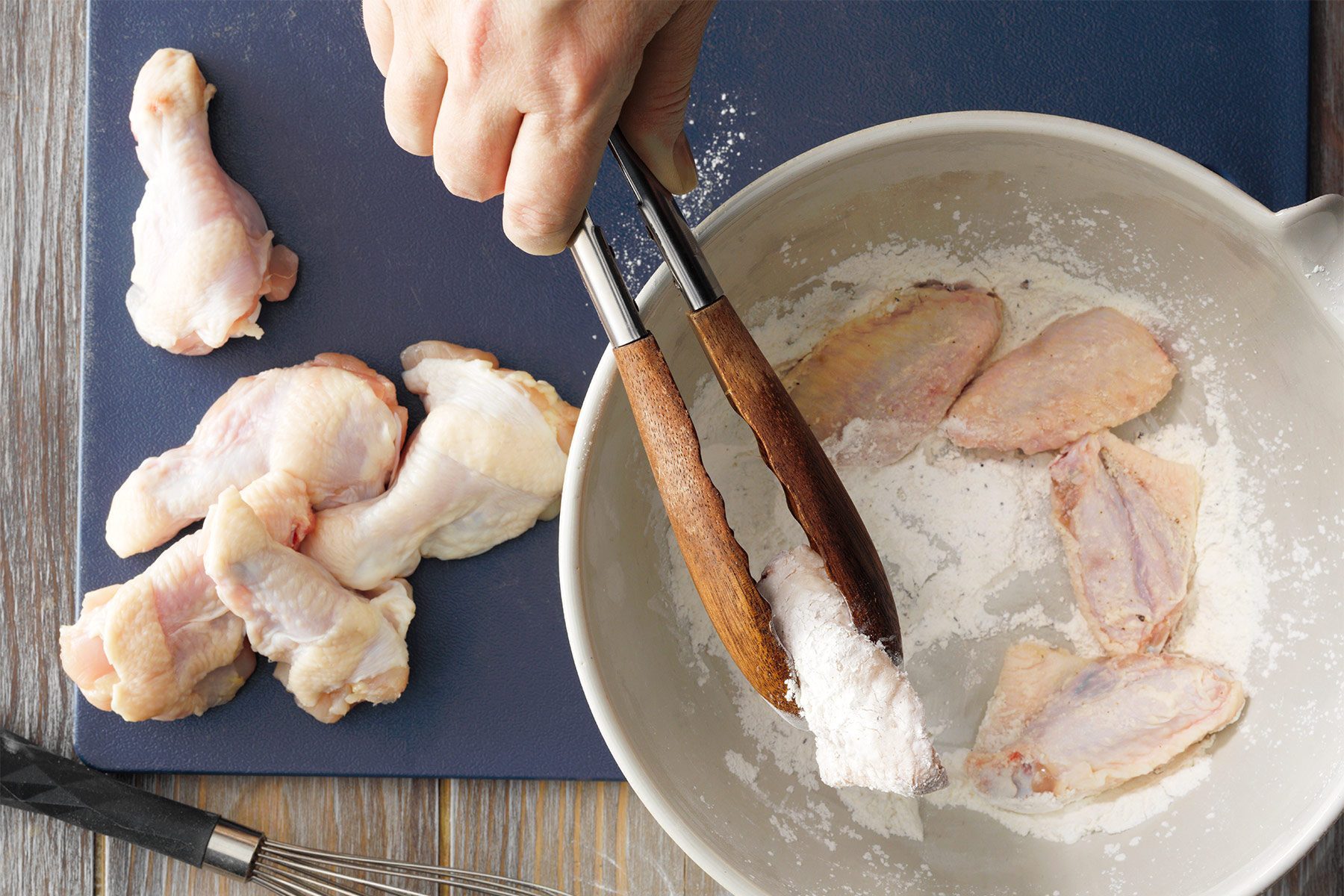 A hand using wooden tongs to coat raw chicken pieces in flour in a white bowl. Nearby, there are more raw chicken pieces on a dark blue cutting board. A metal whisk is partially visible at the bottom left corner of the image.