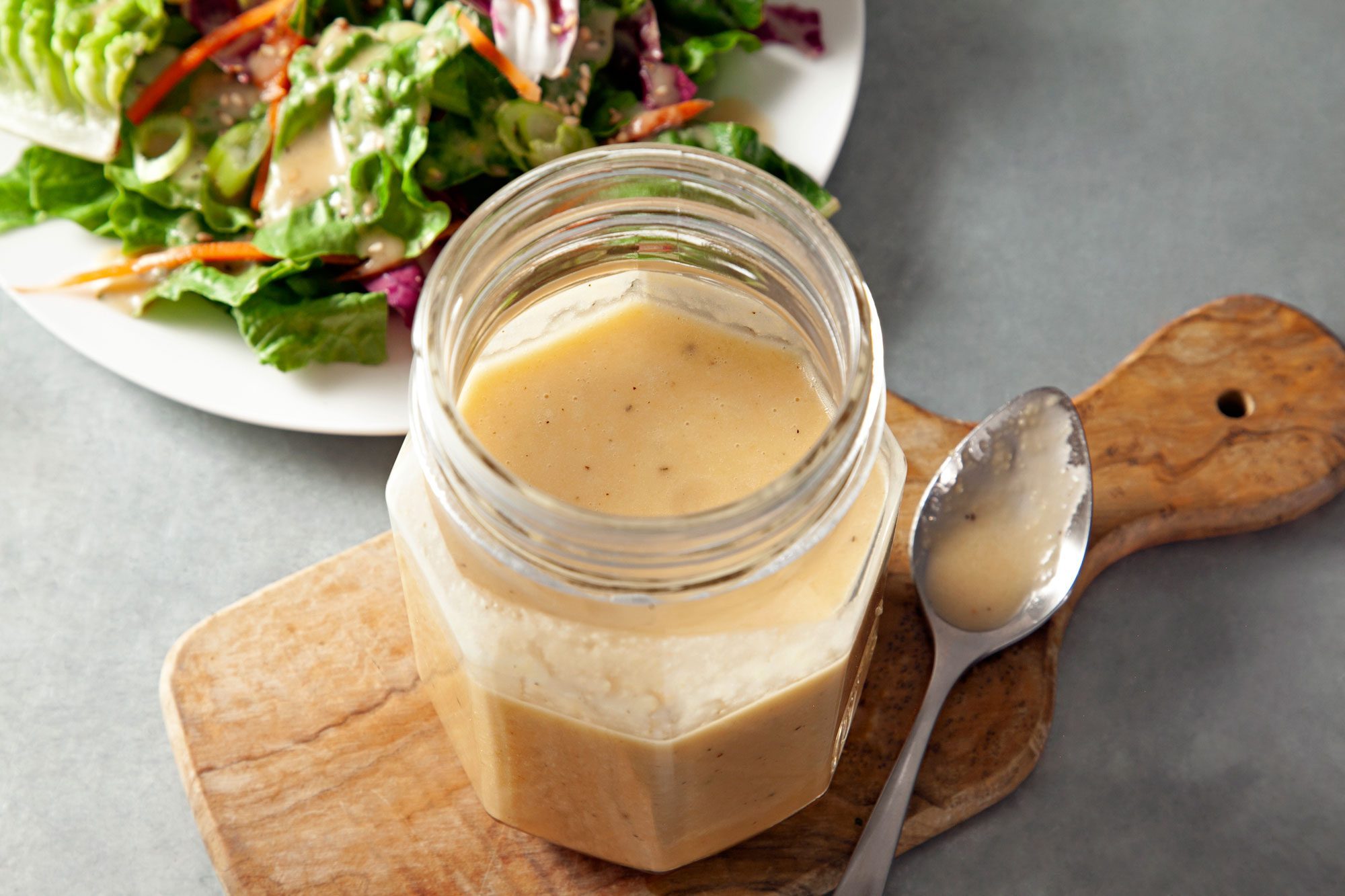 Ginger Dressing in a glass jar placed over wooden board with small spoon; salad in a small white plate ginger dressing spread over it