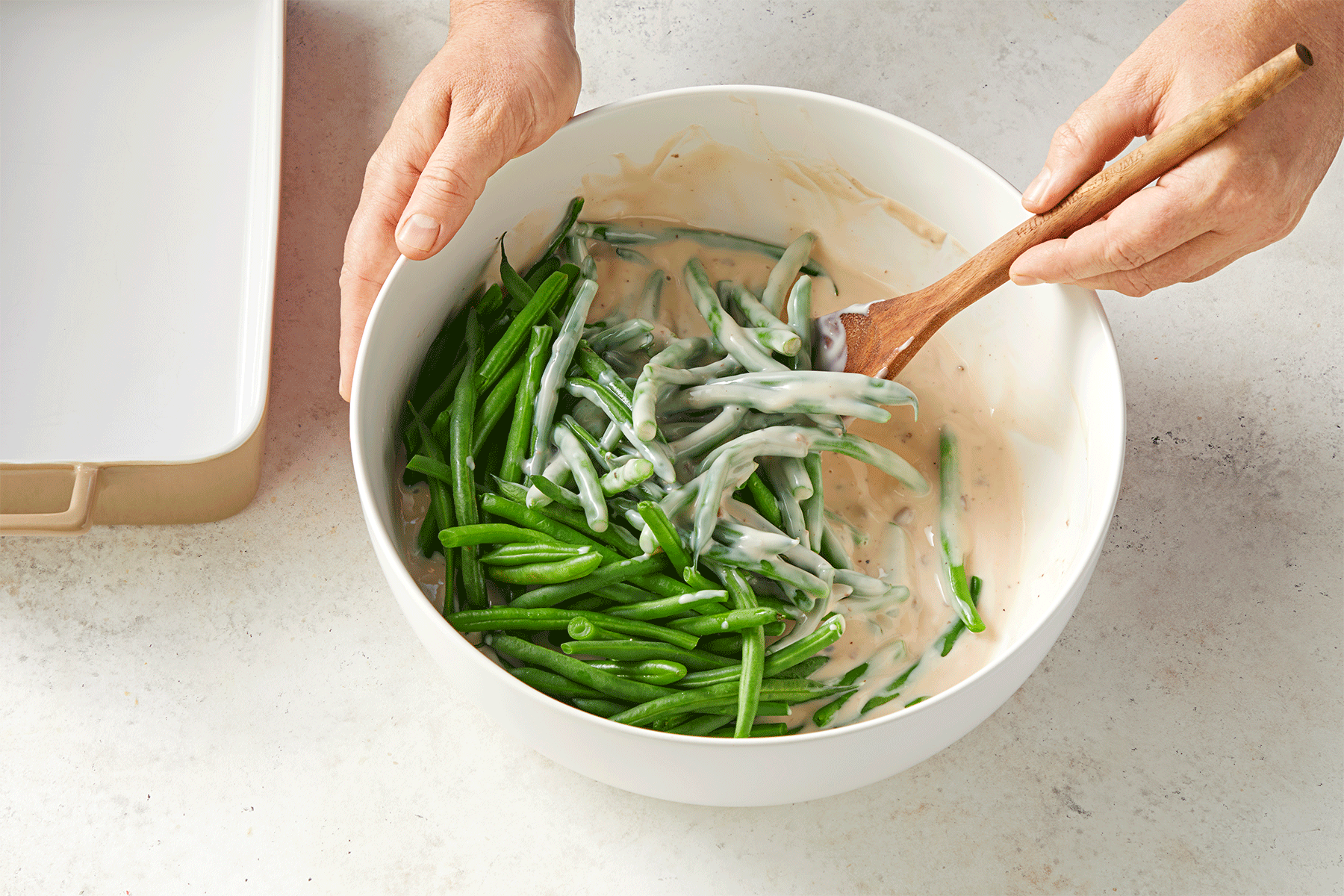 A person is mixing fresh green beans with a creamy sauce using a wooden spoon in a large white bowl.