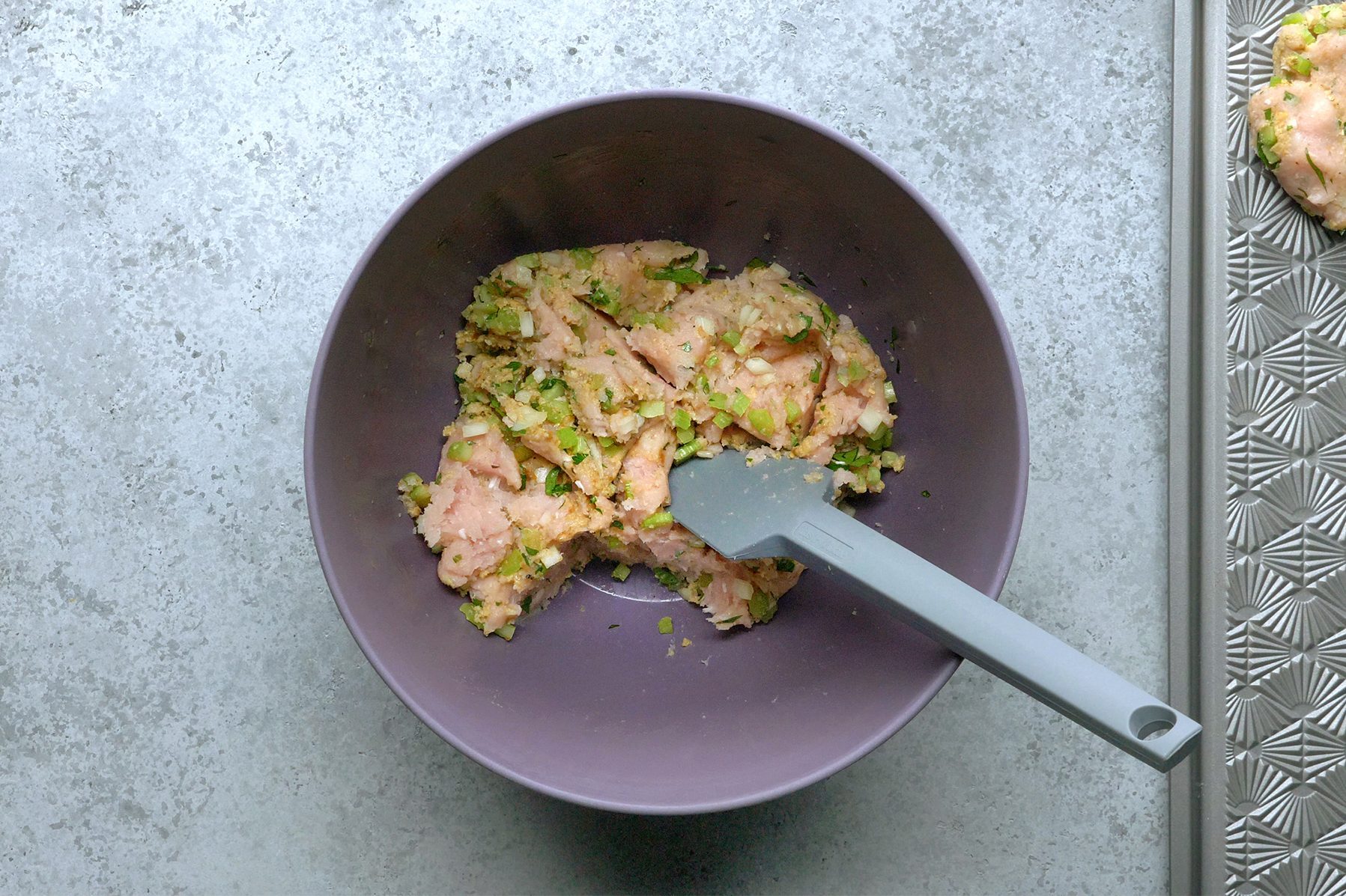 A gray bowl containing a mixture of raw chicken and green vegetables sits on a textured light gray countertop.