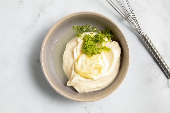 A beige bowl filled with creamy mayonnaise garnished with a sprinkling of lime zest on top sits on a white marble surface.