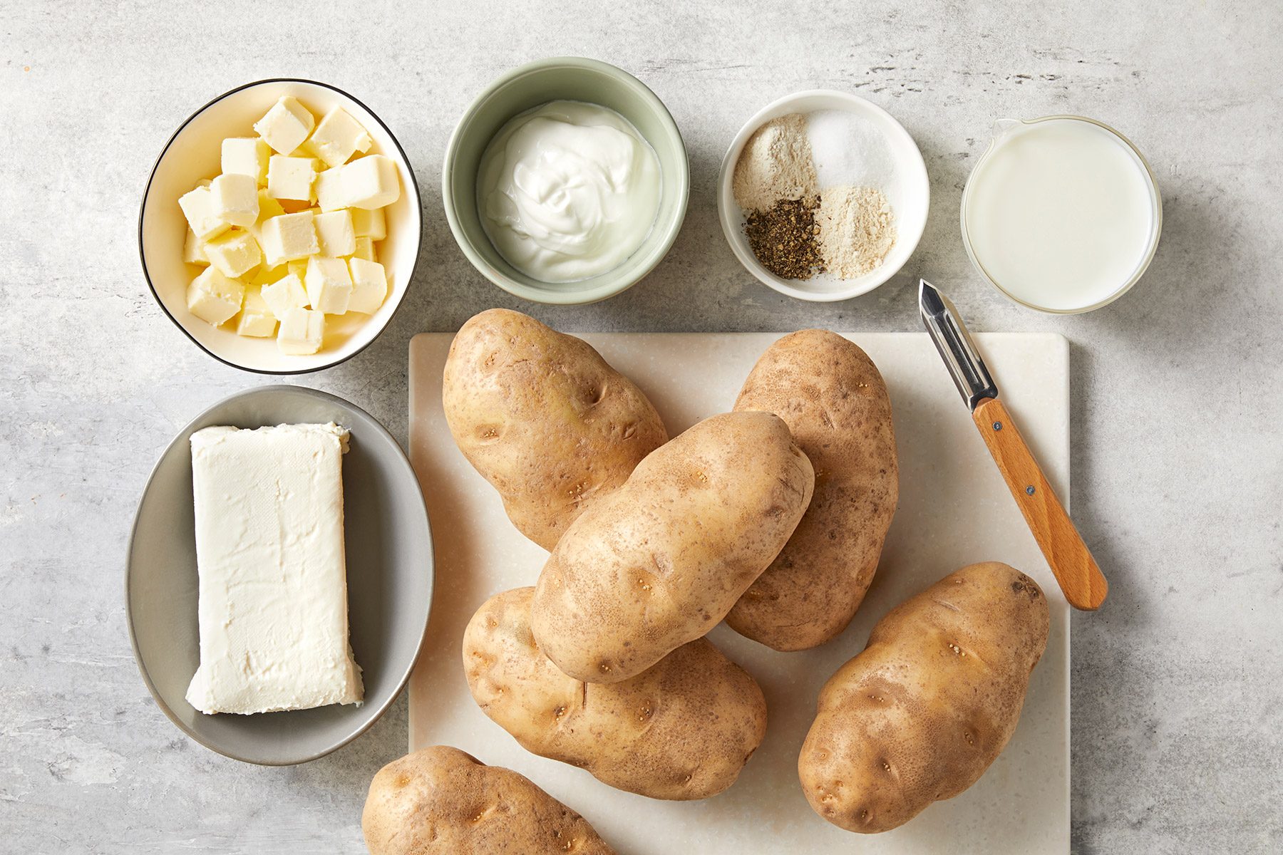 Ingredients for mashed potatoes are arranged on a light surface. Items include several whole potatoes, a bowl of cubed butter, a bowl of sour cream, a bowl of white spices, a bowl of milk, and a block of cream cheese. A peeler lies next to the ingredients.