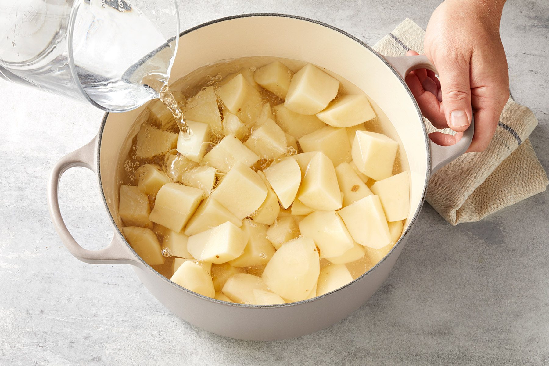 A pot filled with peeled and chopped potatoes placed on a countertop. A person's hand holds the pot handle while water is being poured into the pot, submerging the potato pieces. A cloth lies next to the pot.