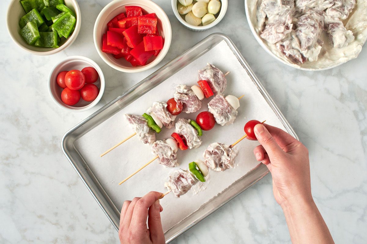 hands assembling pork kabob skewers with bowls of each ingredient nearby
