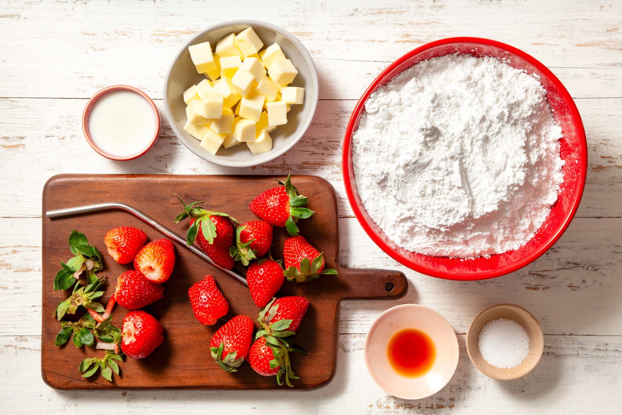 overhead shot of strawberries on wooden chopping board with steel straw; butter in a bowl; all purpose flour in large bowl; white wooden background;
