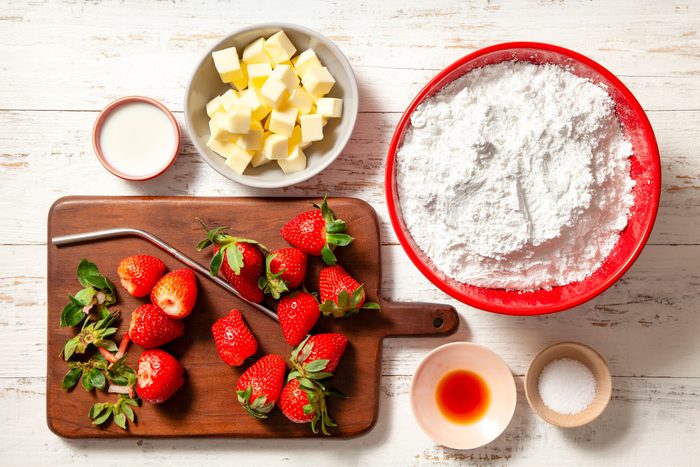overhead shot of strawberries on wooden chopping board with steel straw; butter in a bowl; all purpose flour in large bowl; white wooden background;