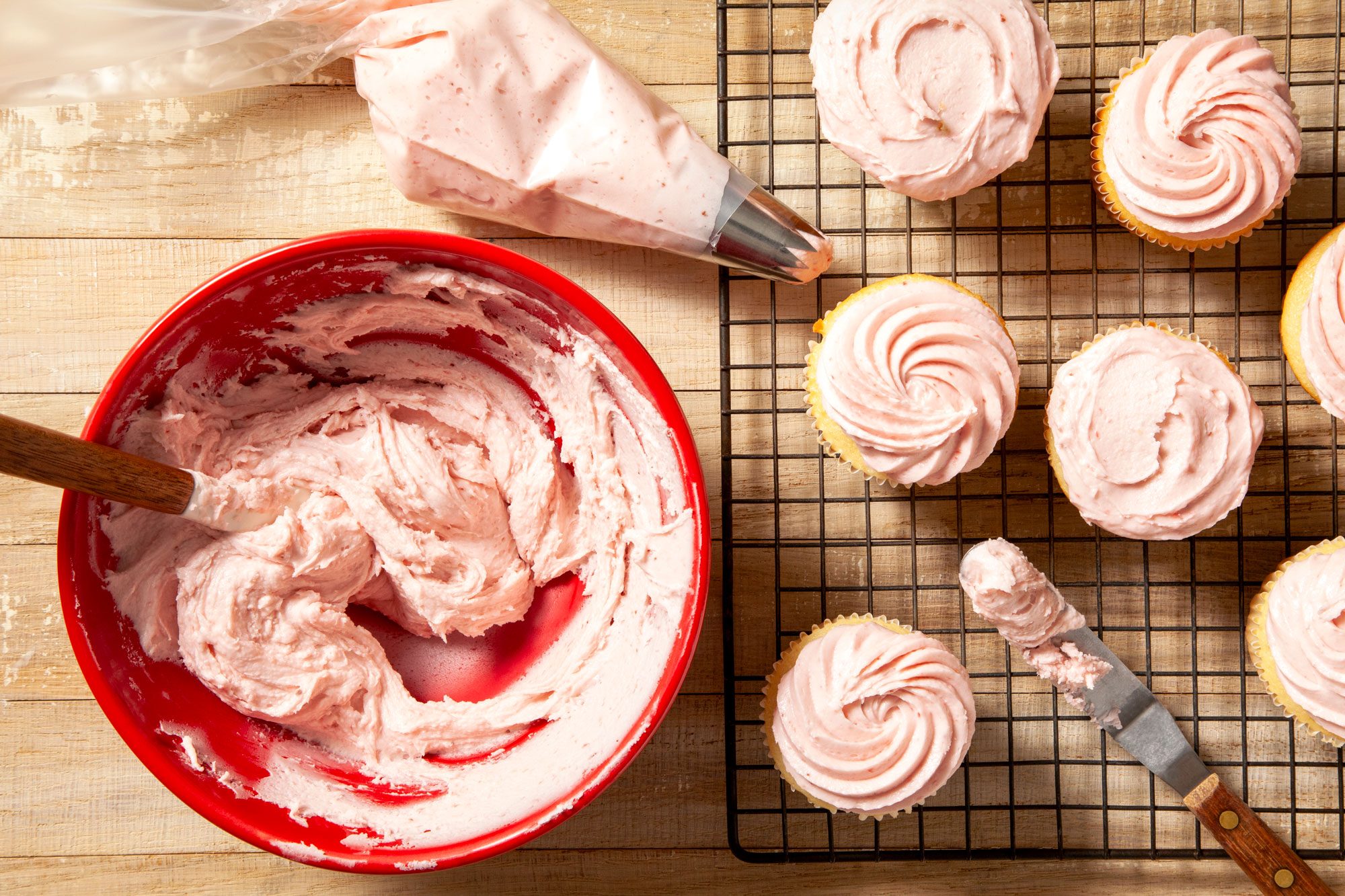 overhead shot of strawberry frosting in large red bowl and knife placed on right side and cup cakes on wired rack on wooden background