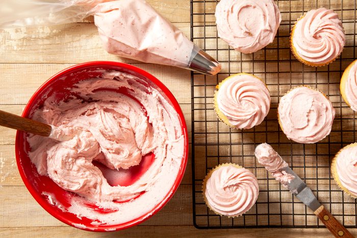 overhead shot of strawberry frosting in large red bowl and knife placed on right side and cup cakes on wired rack on wooden background