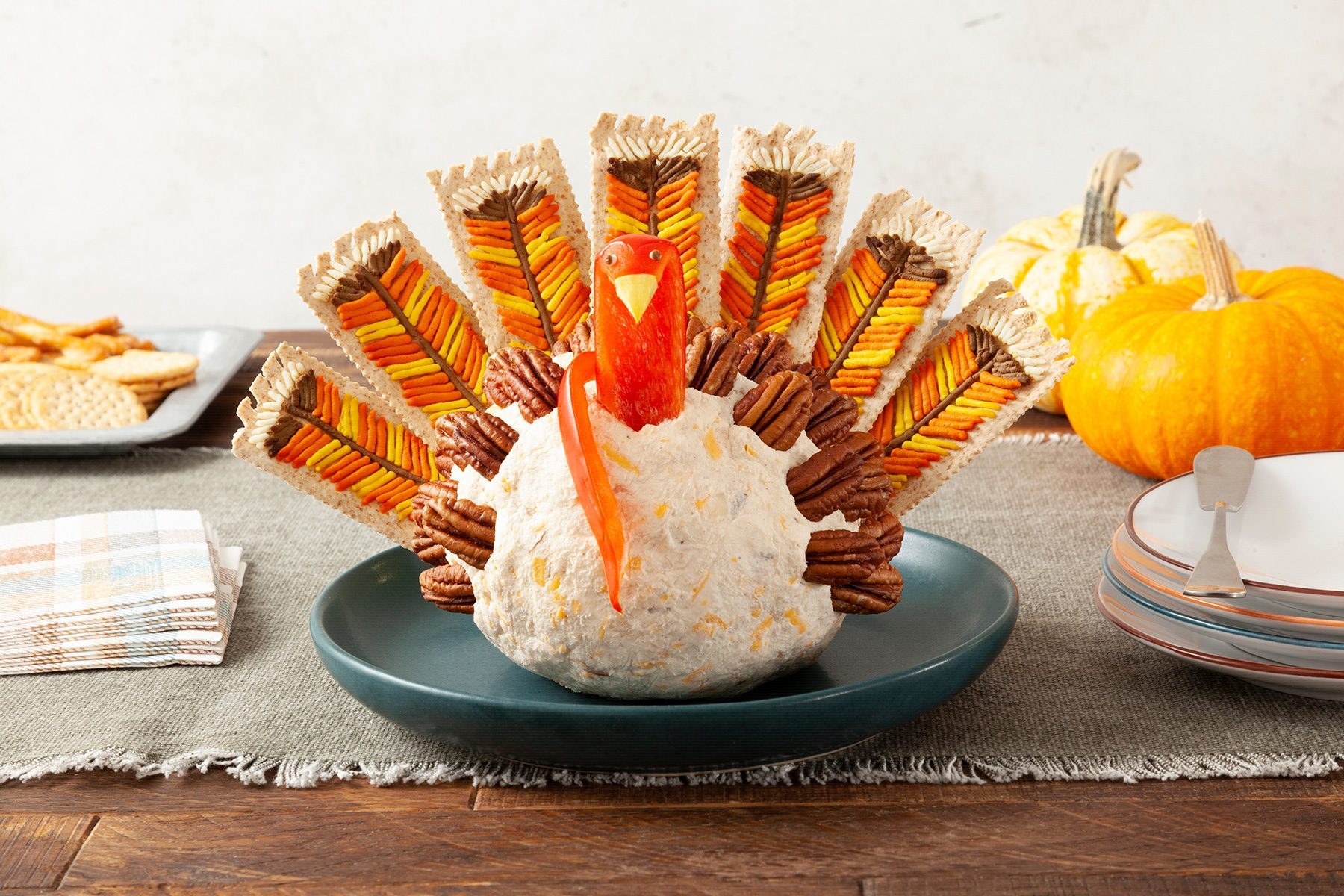 A cheese ball appetizer styled as a turkey sits on a green plate. The turkey's body is a round cheese ball, its feathers are colored crackers, and its wings are pecans. A red pepper forms the turkey's head and beak.
