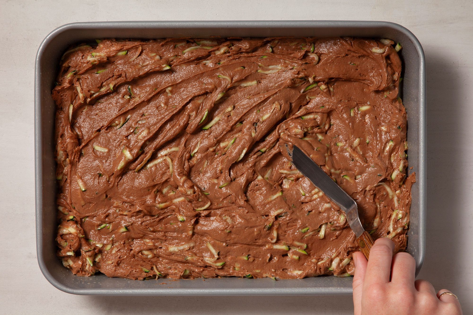 Overhead shot of pour Zucchini Brownies mix into a greased baking pan; cake knife; marble background;