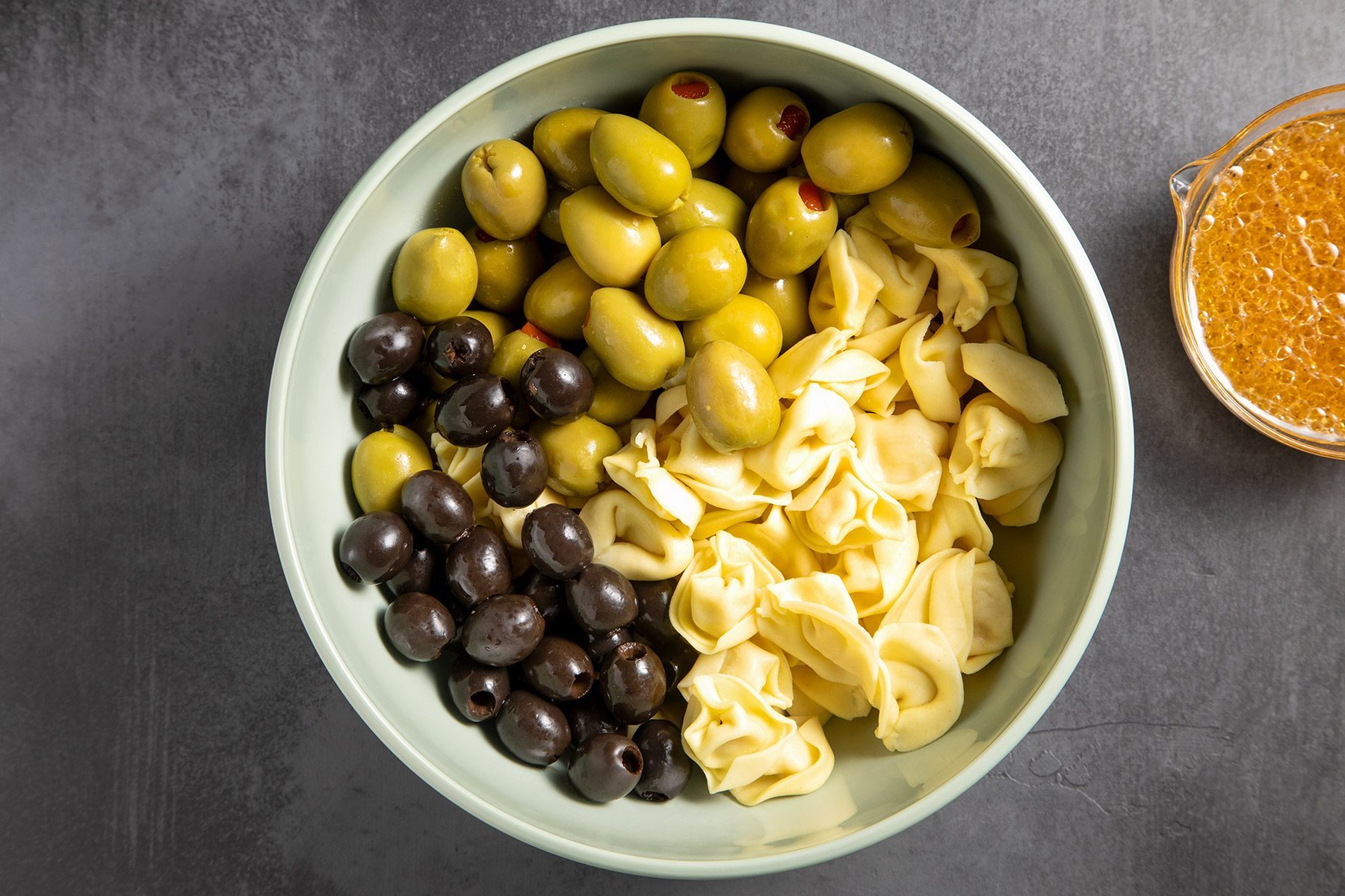 overhead shot; grey textured background; In a large bowl, combine the tortellini, olives and salad dressing in a small bowl