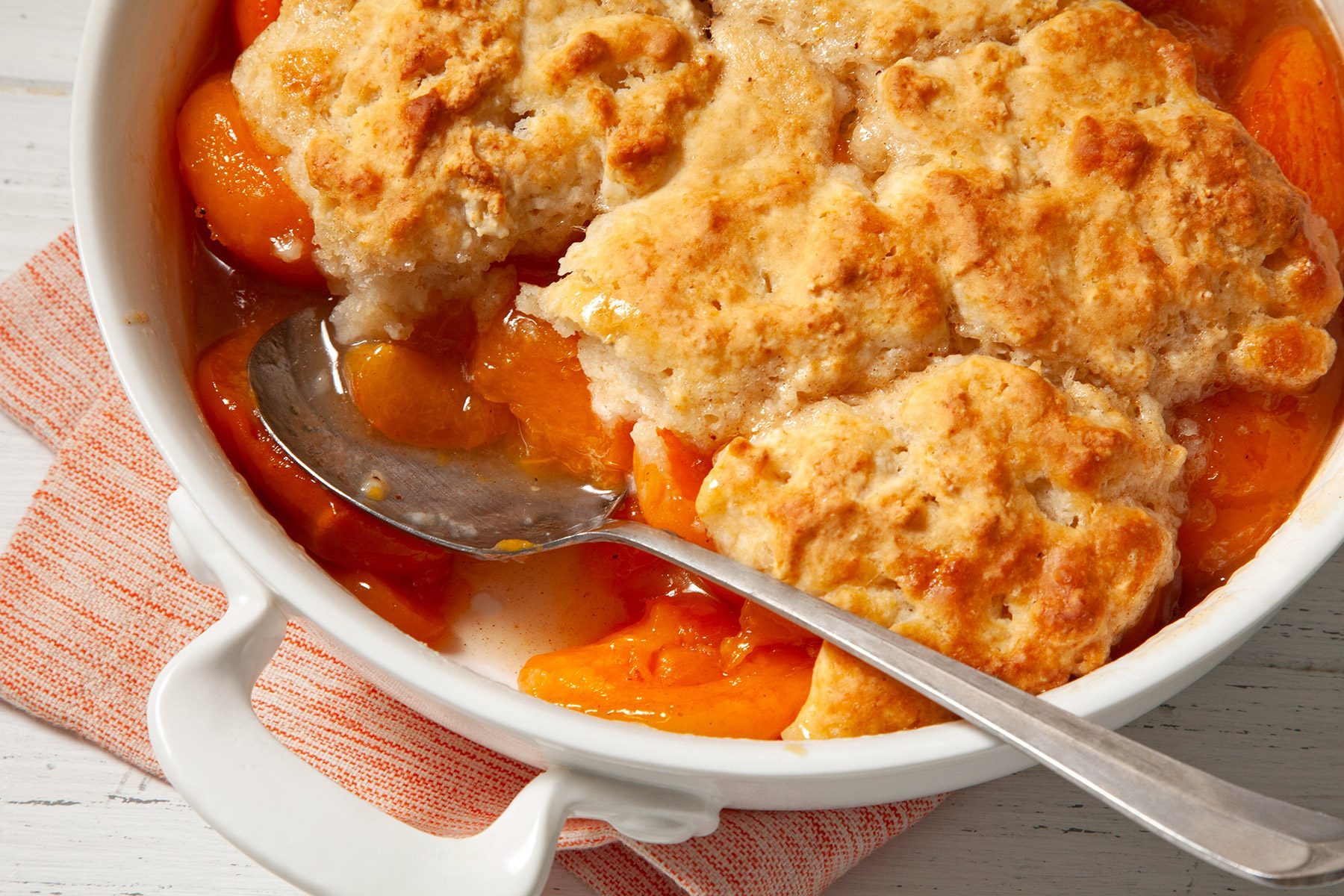 3/4th shot; white background; Apricot Cobbler in a large white dish with large silver spoon over kitchen towel