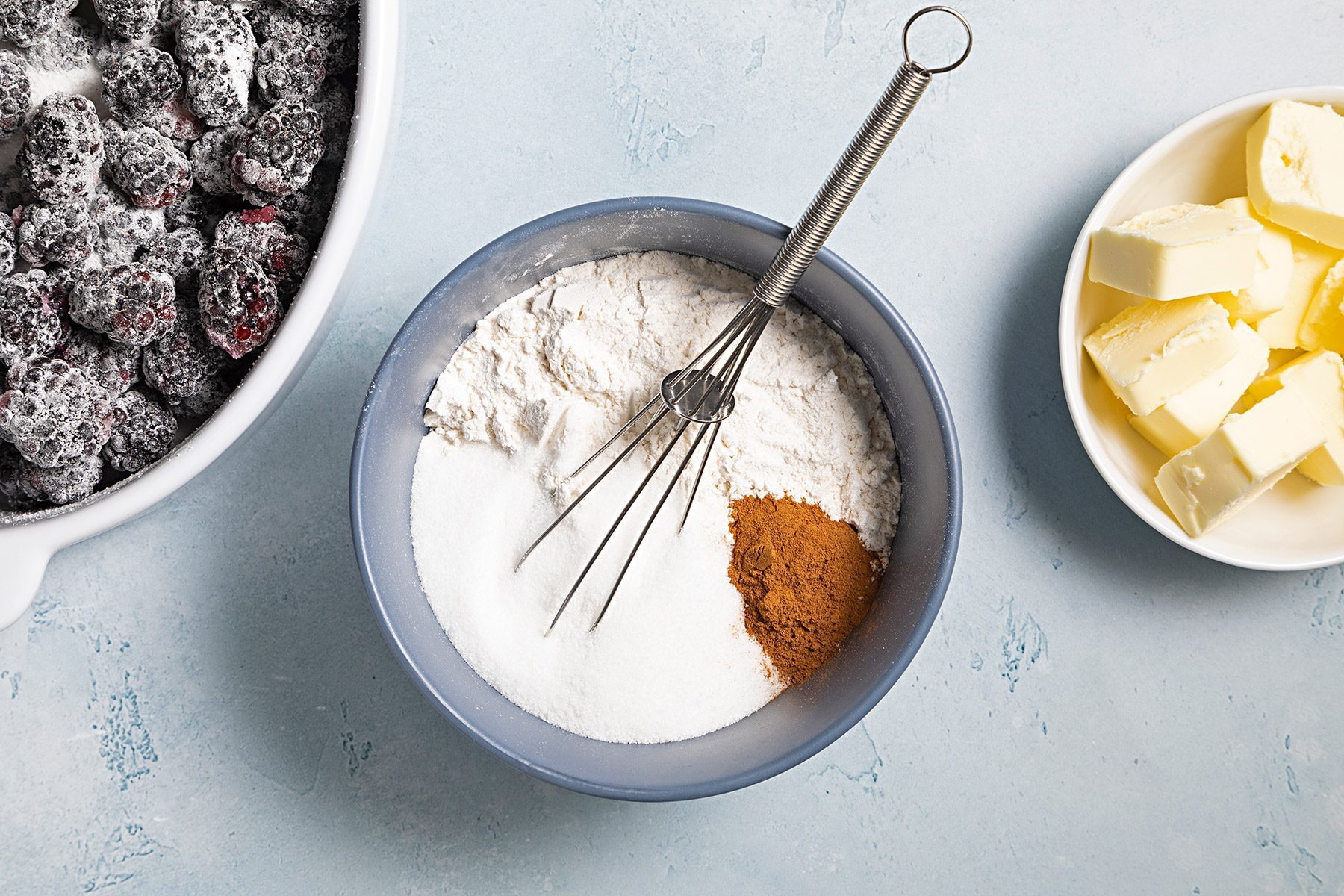 Overhead shot of combine flour; sugar; cinnamon and salt; mix well; whisk tool; butter bowl; coated blackberries bowl; blue marble background