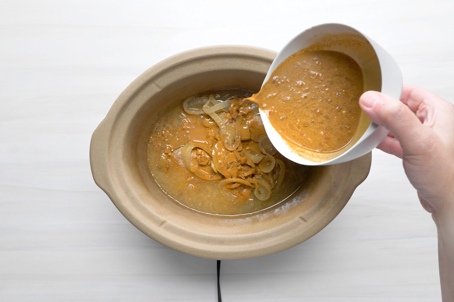 A hand is pouring gravy over beef cube steaks in a slow cooker