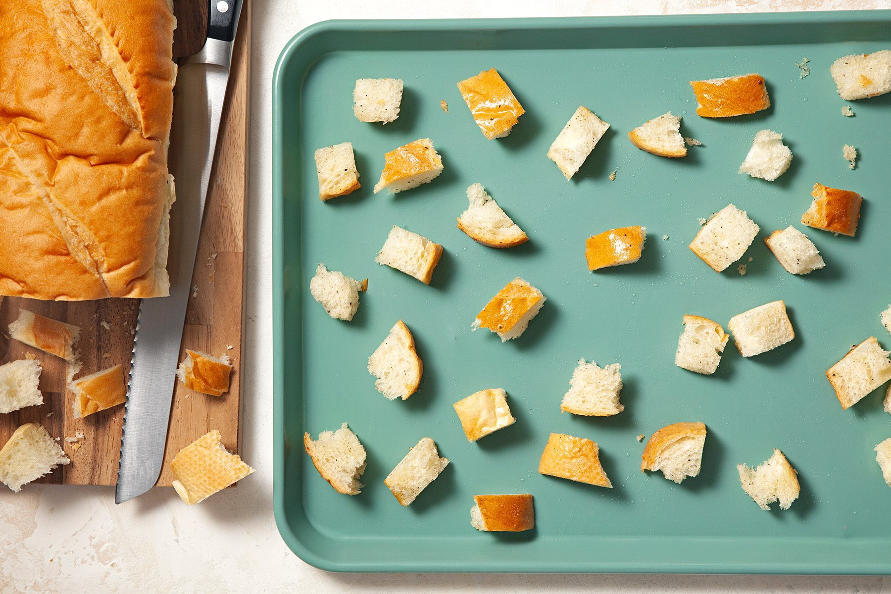 Overhead shot of large bread on chopping board; cut in small pieces; place in baking pan; knife; cream marble background