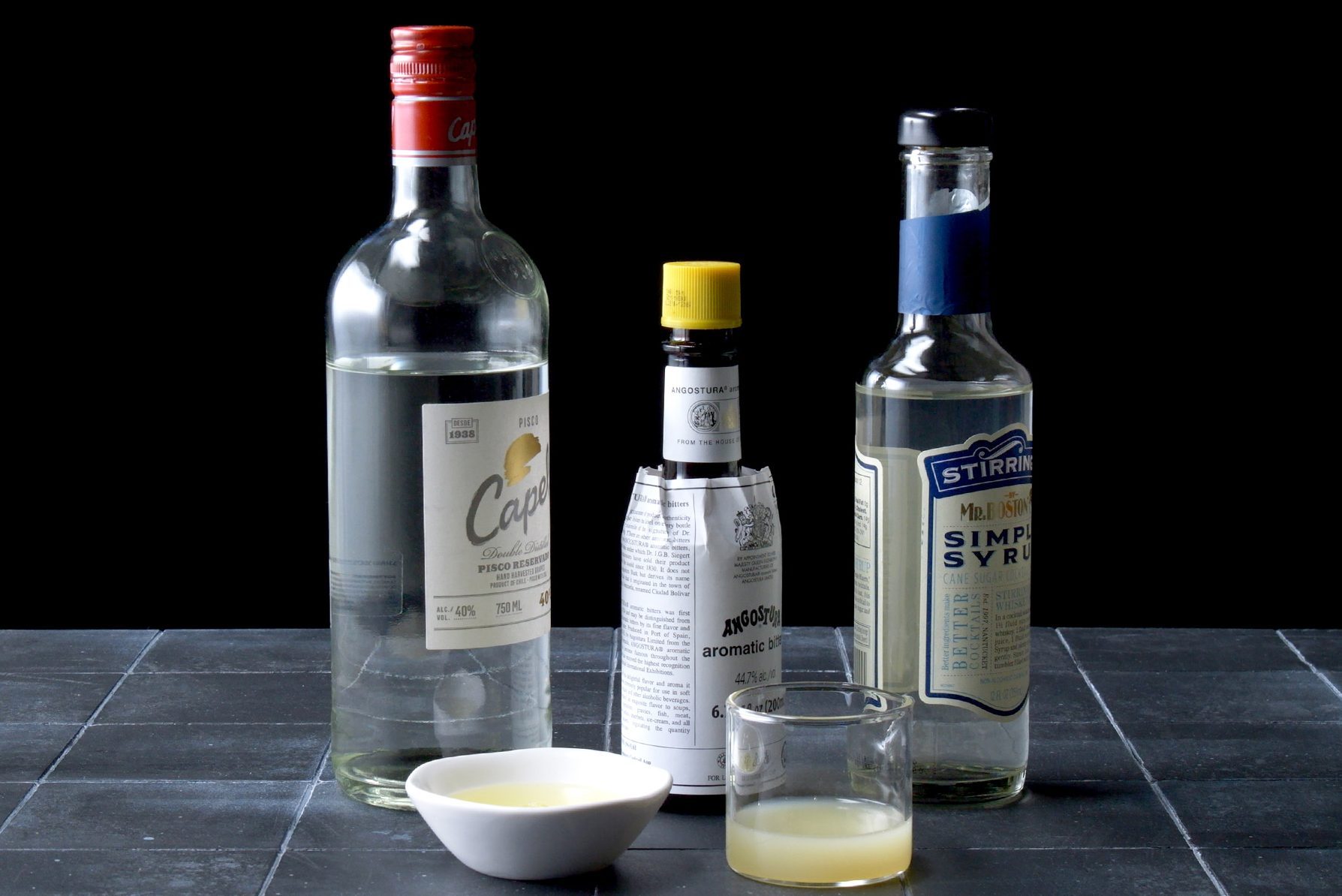 A collection of drink ingredients on a dark surface, featuring a large clear bottle with a red cap, a small bottle with a yellow cap, a bottle of simple syrup, a white bowl with liquid, and a glass with a pale yellow liquid.