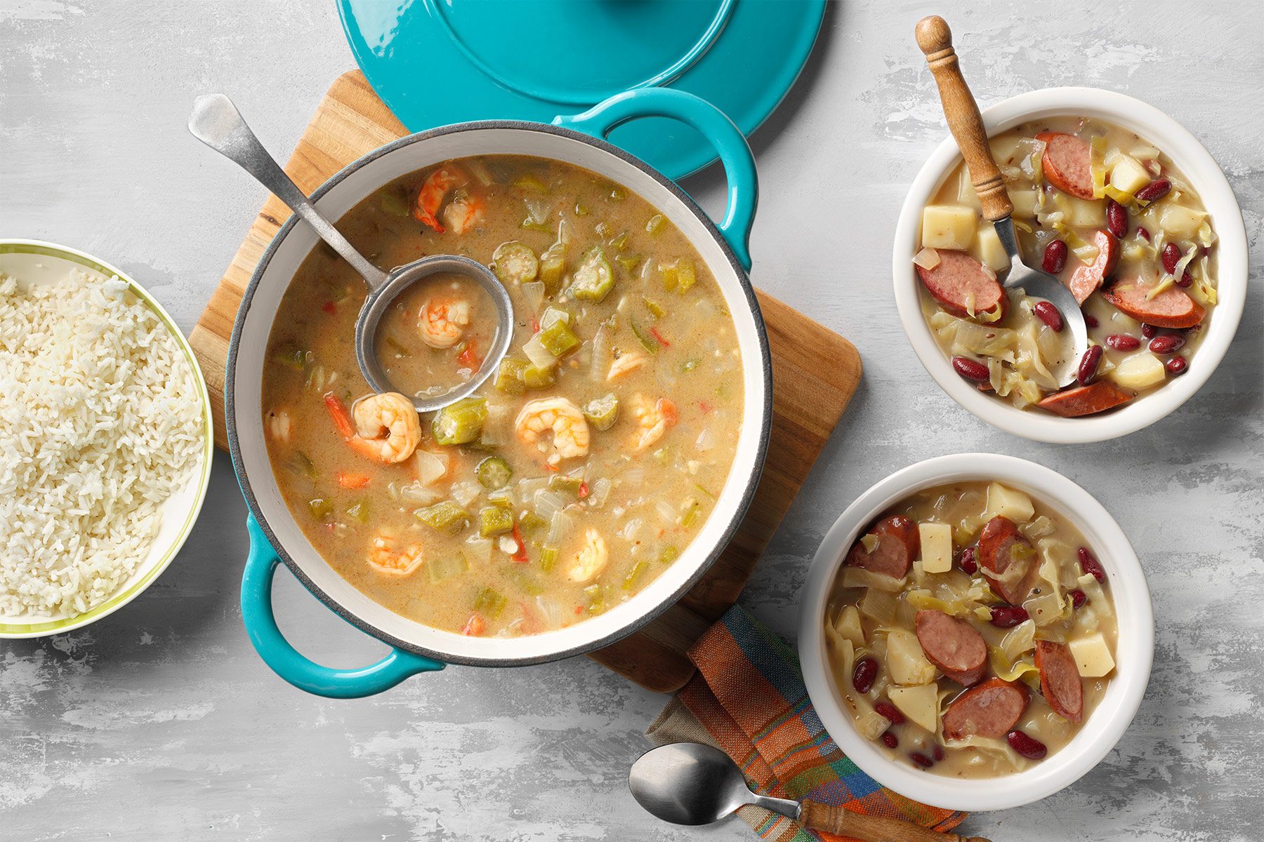 A pot of hearty seafood stew with ladle on a wooden board. The stew contains shrimp, vegetables, and sausage. Two bowls of the same stew accompanied by spoons are placed beside the pot. A striped cloth is partially visible under the bowls.