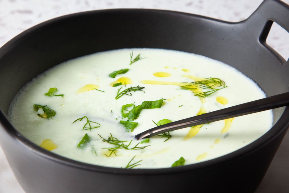 Single servings of Taste of Home Cucumber Soup in black bowls on a light gray plates, topped with olive oil, green onions and dill