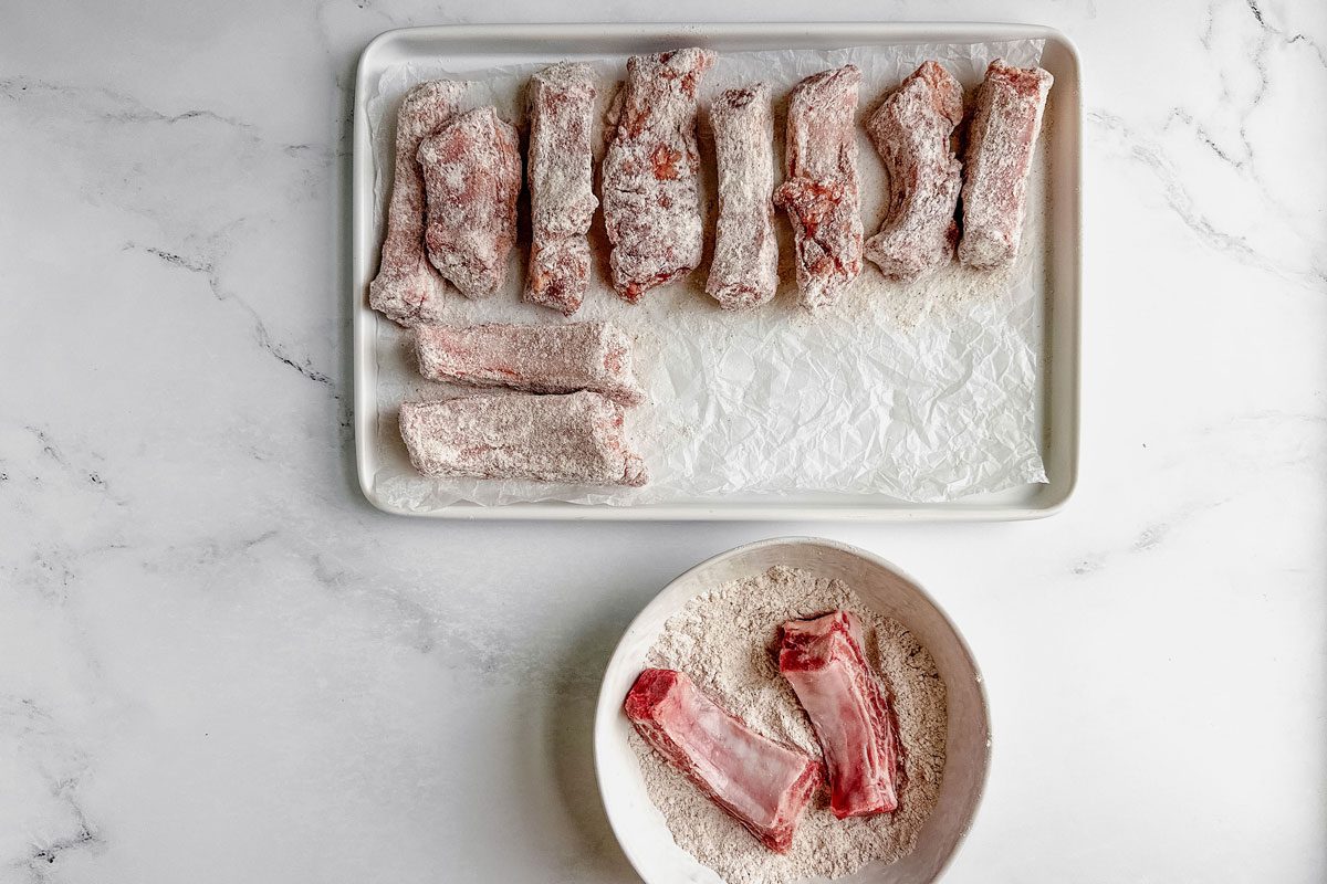 Floured ribs for Taste of Home fried ribs on a white tray and in a white bowl on a marble surface.