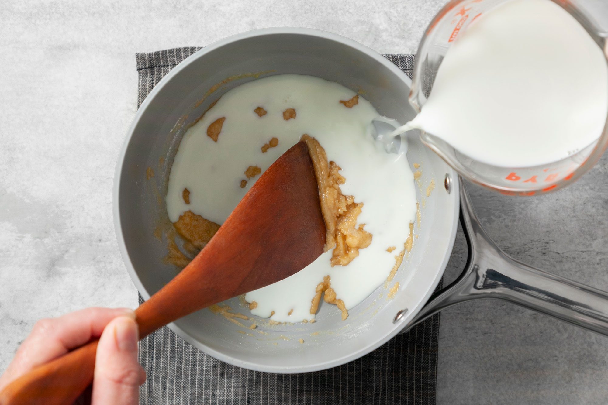 Overhead shot of same saucepan; gradually whisk in milk; bring to a boil; stirring constantly; cook and stir until thickened; wooden spatula; kitchen cloth; grey marble background;
