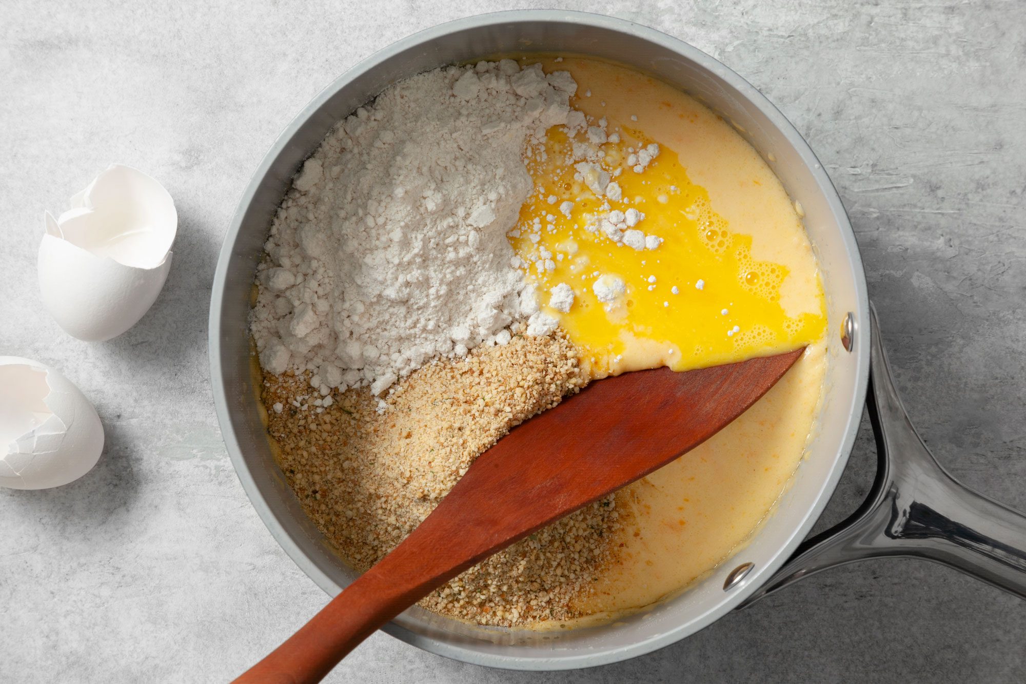 Overhead shot of same saucepan; remove from heat; stir in biscuit mix; eggs and 1/2 cup bread crumbs; wooden spatula; grey marble background;