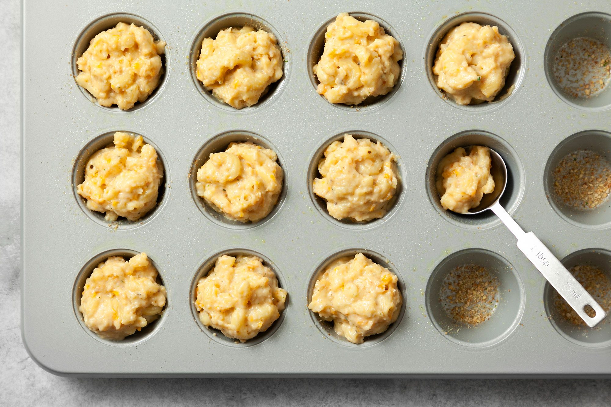 Overhead shot of mini muffic cups baking tray; spoon about 2 tablespoons macaroni mixture into each prepared mini-muffin cups; grey marble background;