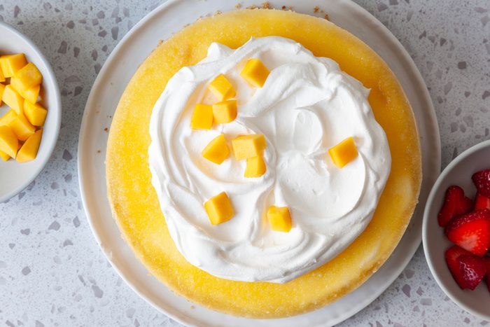 Step 5 of Taste of Home Mango Cheesecake is to garnish the cheesecake with whipped cream and additional mango and strawberries