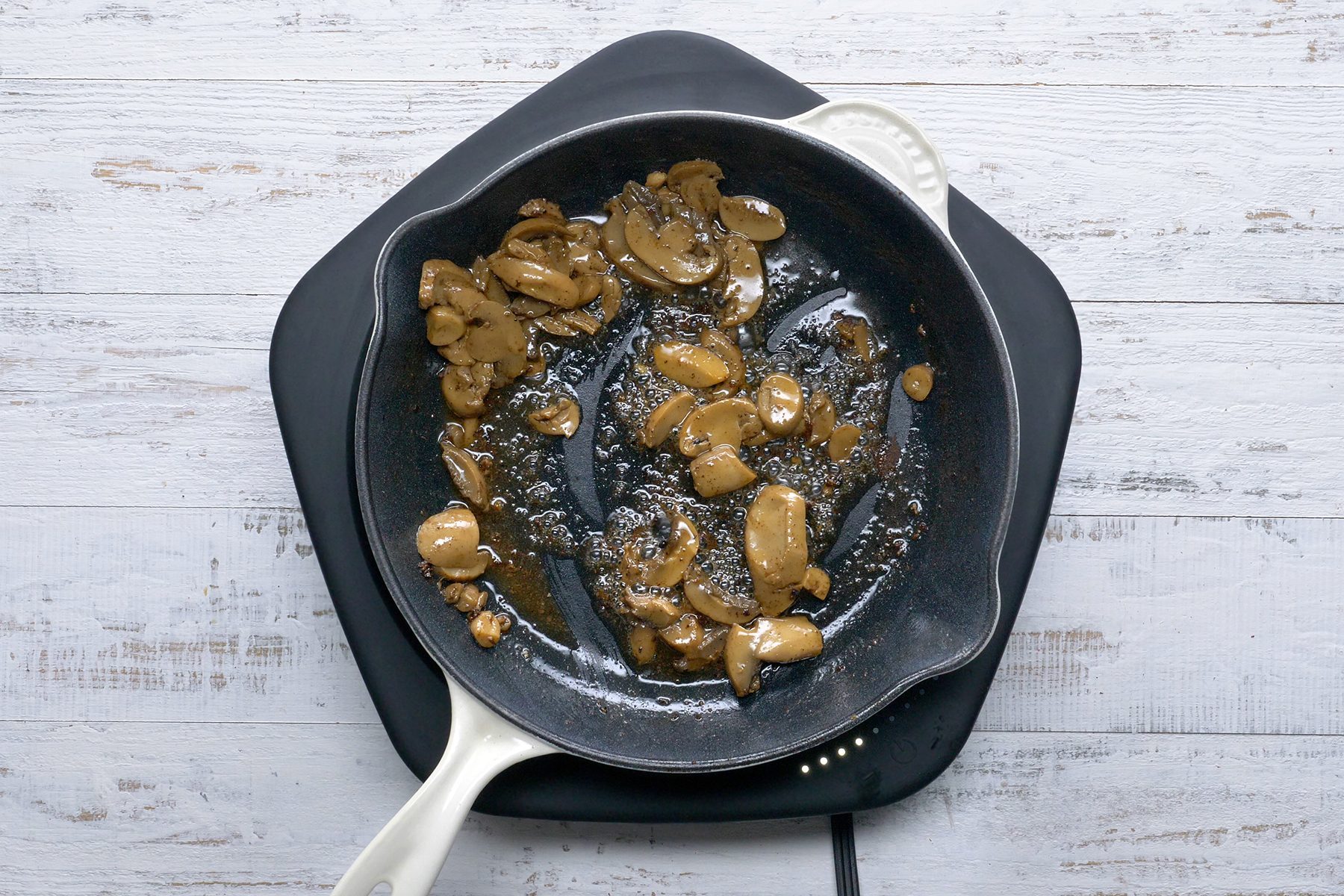 A white-handled frying pan on an electric stove contains sautéed sliced mushrooms. The mushrooms are light brown and glistening with oil, sitting on a dark surface. 