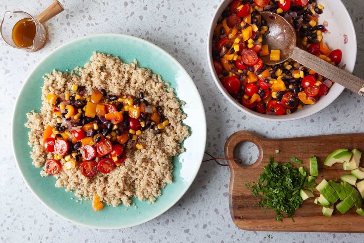 Large serving bowl with quinoa being topped with vegetable mix and a cutting board with cilantro and avocado on the surface