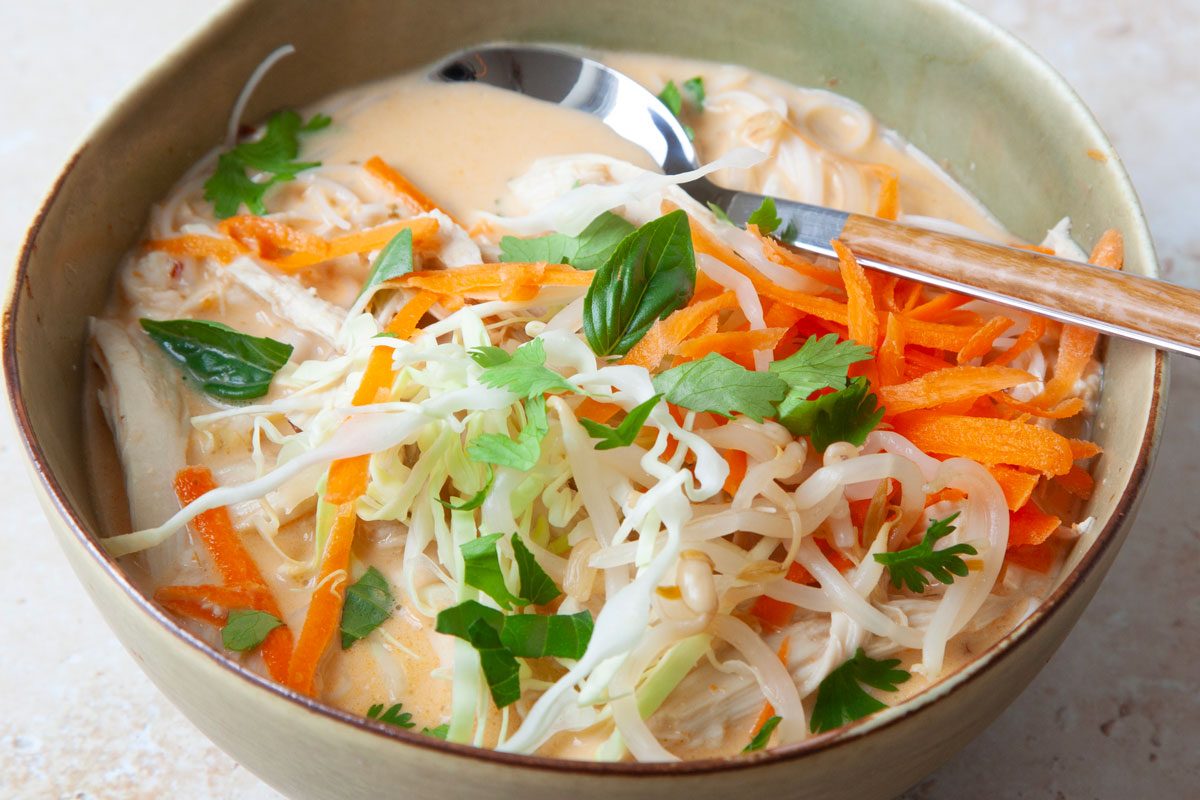 Coconut curry chicken soup in green bouls with carrot and cabbage garnish, basil and cilantro on a cutting board,