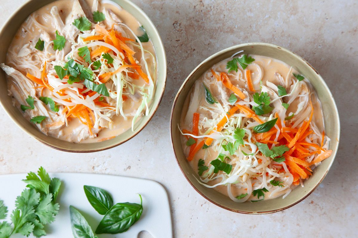 Coconut curry chicken soup in green bouls with carrot and cabbage garnish, basil and cilantro on a cutting board,