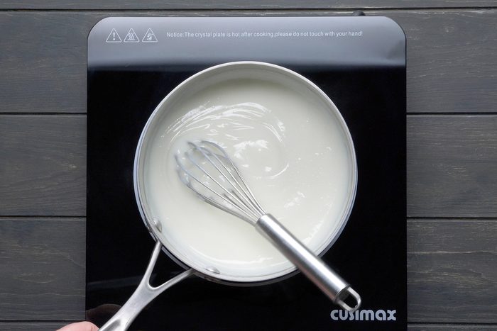 A saucepan sits on an induction stove, containing a white liquid being stirred with a whisk. The induction stove has the brand name "Cusimax" on it and a warning message indicating that the crystal plate is hot after cooking and should not be touched by hand.