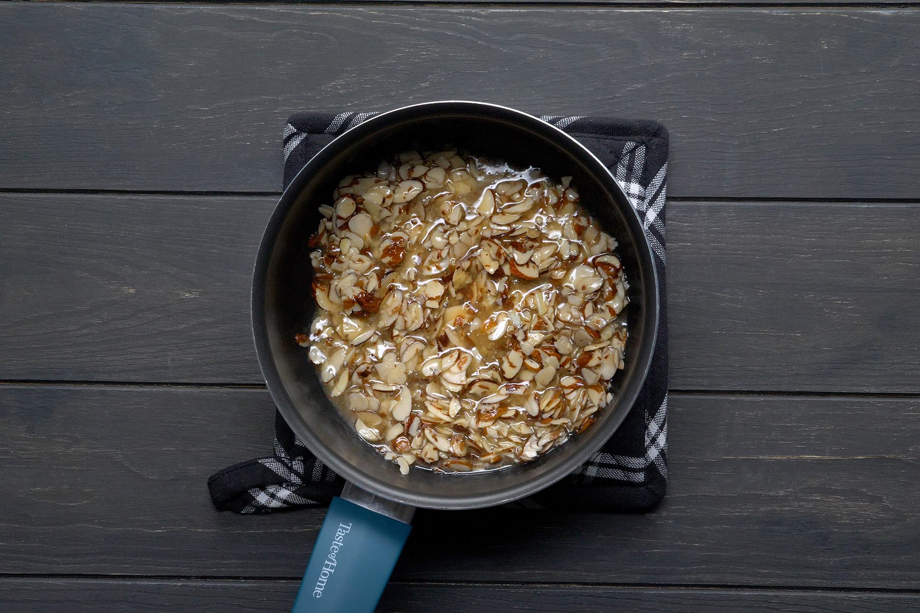 A saucepan containing sliced almonds being cooked in oil is placed on a dark, textured wooden surface. 