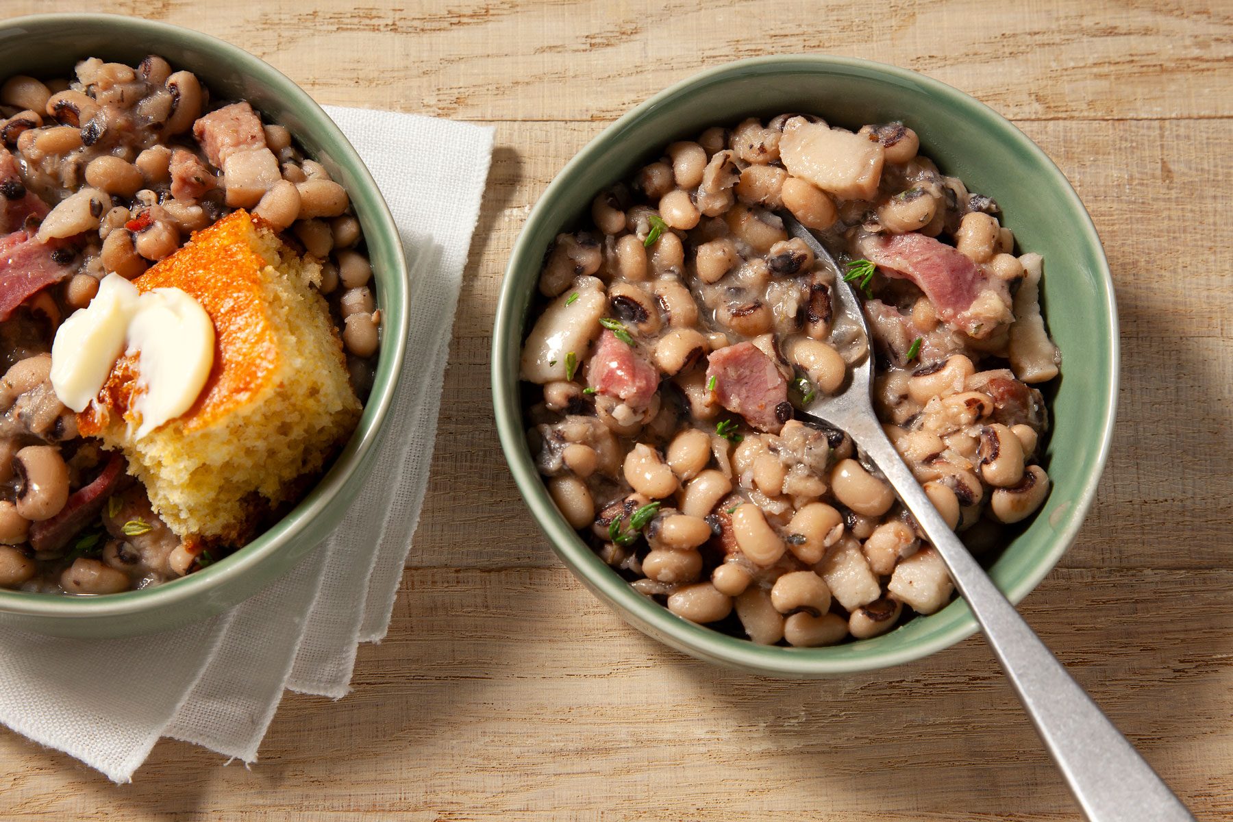 Two green bowls filled with black-eyed peas, chopped meat, and herbs on a light wooden surface. One bowl also contains a piece of cornbread topped with butter pats. A white napkin is partially visible under the bowl with cornbread.