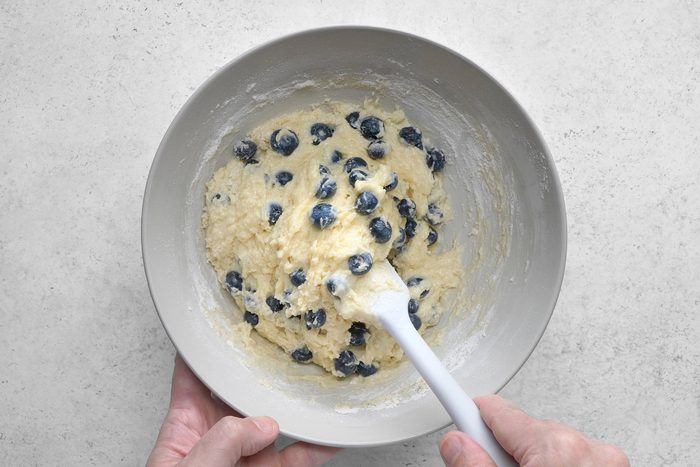 A bowl containing a thick batter mixed with blueberries is being stirred with a white spatula. The bowl is being held by two hands over a light gray countertop.