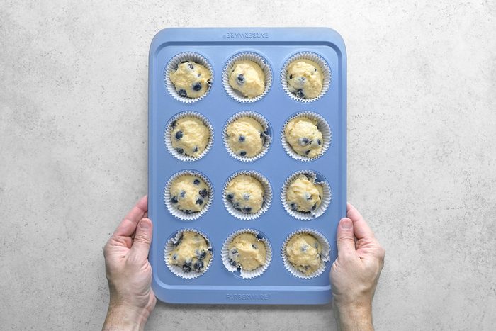 Two hands holding a blue muffin tray with twelve compartments filled with blueberry muffin batter in white paper liners, ready to be baked. The background is a light, textured surface.