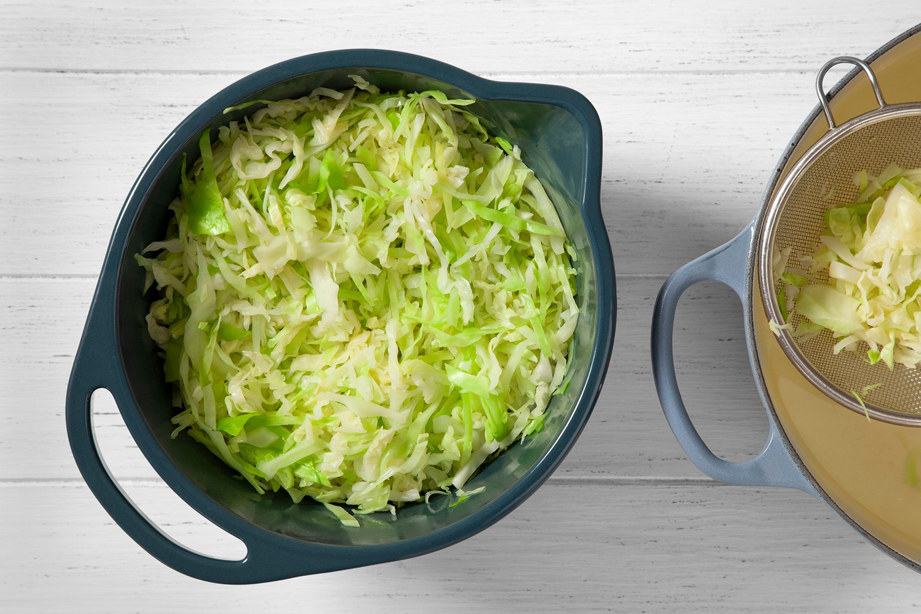 A blue colander filled with shredded green cabbage is on a white wooden surface. To the right, a small amount of shredded cabbage is captured in a metal strainer inside another blue bowl.